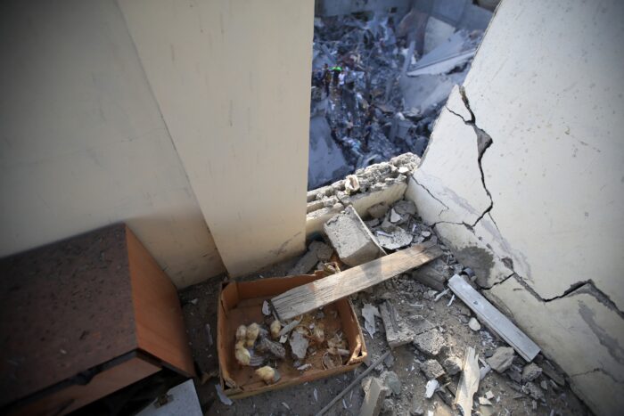 Blick durch eine geborstene Mauer in einem höheren Stockwerk, unten sind in der Ferne Menschen zu sehen, die in Trümmern etwas suchen