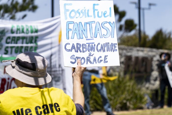 Ein Mann von hinten in gelber Weste hält ein Schild auf einer Protestaktion hoch, auf dem steht: Fossil Fuel Fantasy = Carbon Storage & Capture.