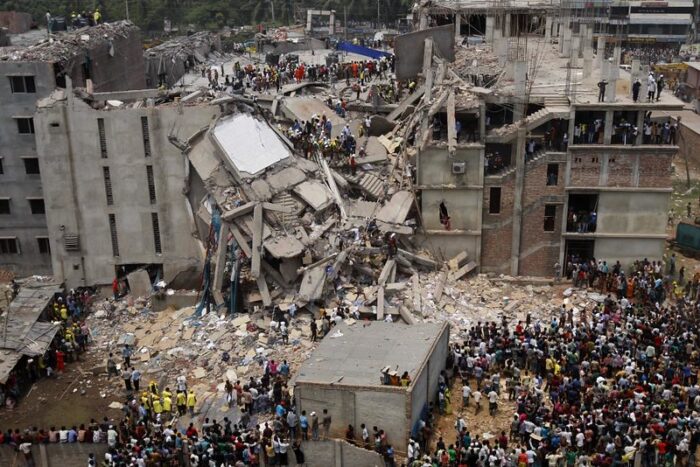 Auf dem Foto ist das eingestürzte Fabrikgebäude der Textilfabrik Rana Plaza in Bangladesh zu sehen. Das Bild ist von einem erhöhten Punkt aufgenommen. Etliche Menschen stehen vor dem Gebäude und sehen sich den Schaden an. Auch auf dem kaputten Gebäude sind Menschen unterwegs, viele in Schutzausrüstung