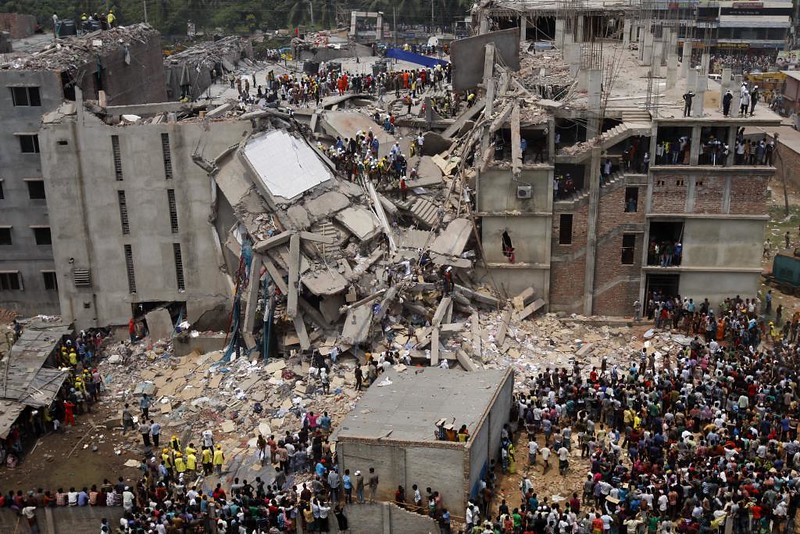 Auf dem Foto ist das eingestürzte Fabrikgebäude der Textilfabrik Rana Plaza in Bangladesh zu sehen. Das Bild ist von einem erhöhten Punkt aufgenommen. Etliche Menschen stehen vor dem Gebäude und sehen sich den Schaden an. Auch auf dem kaputten Gebäude sind Menschen unterwegs, viele in Schutzausrüstung
