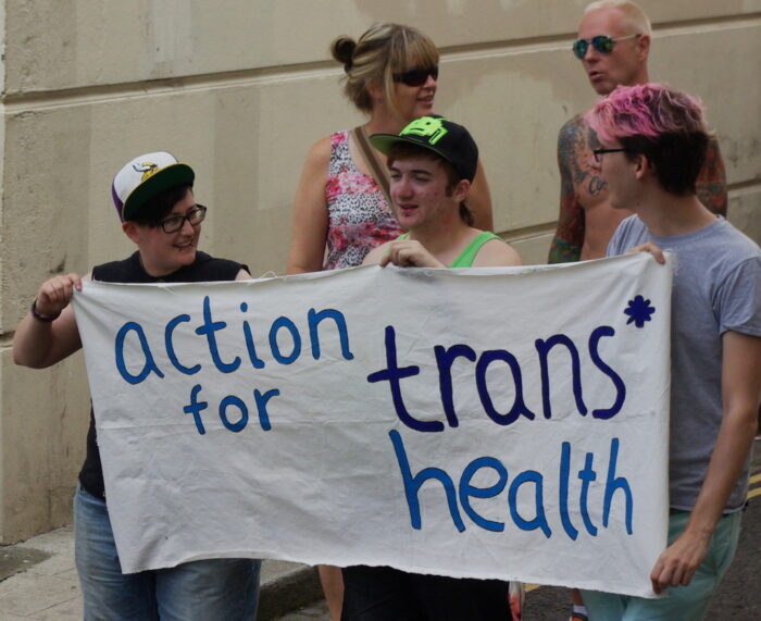 Foto von drei Personen die ein Transpi halten. Auf dem Transpi steht "action for trans health"