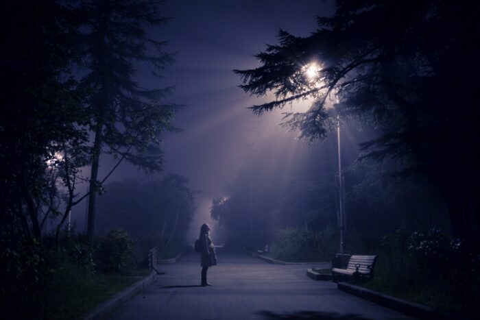 Dunkle Nacht, eine Frau auf einem Gehweg, die nach oben auf ein Licht schaut, das aussieht wie ein Ufo - oder wie eine Straßenlaterne.