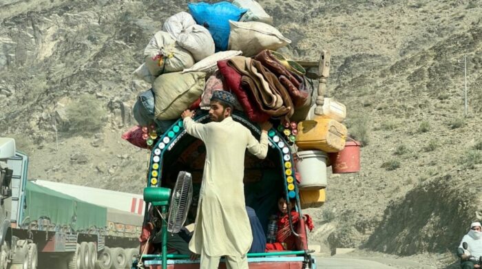 Foto eines Menschen in langen beigen Gewändern der hinter einem vollbeladenen Hänger herläuft und die gestapelten Sachen festhält