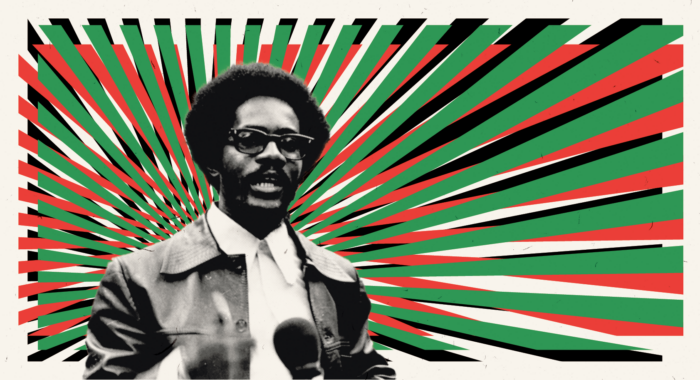 Ein Mann mit Brille und Lederjacke spricht in ein Mikrofon, dahinter aufgefächert die panafrikanischen Farben, grün, rot, schwarz.