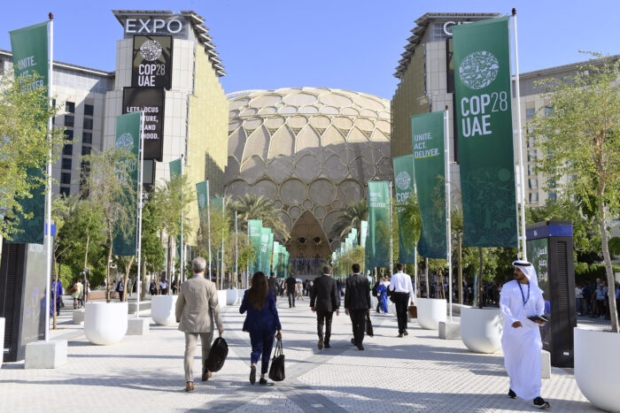 Eingangsbereich mit grünen Flaggen der COP28, auf den vereinzelt Menschen zulaufen.