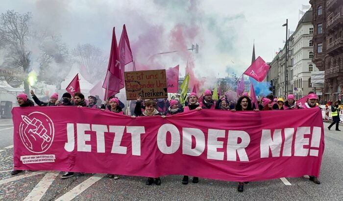 Junge studentische Beschäftigte tragen ein pinkes Banner auf einer Demonstration. Auf dem Banner steht: Jetzt oder nie! Daneben ist das Symbol der Bewegung TVStud zu sehen. Es besteht aus einer Faust, die einen Bleistift hält. Im Hintergrund wehen pinke Fahnen und pinker, sowie blauer Rauch, von Rauchtöpfen steigt auf. In der zweiten Reihe hält jemand ein Schild in die Luft auf dem steht: Es ist uns 1 Ehre an der Uni zu streiken.