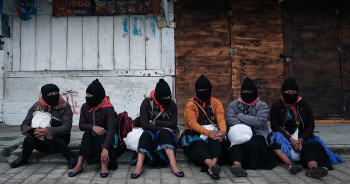Zapatistische Aktivist*innen in Chiapas sitzen am Rande einer Soli-Demonstration für die Ukraine an einem Boardstein. Sie tragen schwarze Strumpfmasken über dem Kopf