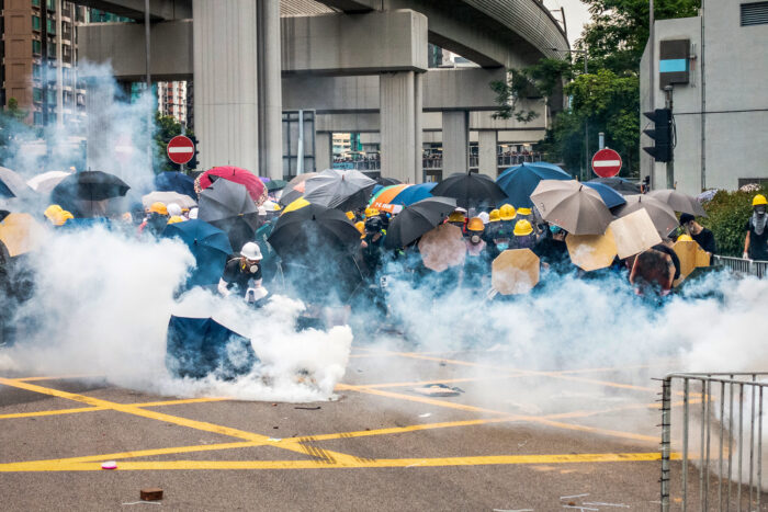 Demonstrierende in Hongkong verschanzen sich hinter Regenschrirmen. Offensichtlich sind Tränengasgranaten explodiert. Einige tragen Gasmasken, man sieht Rauch