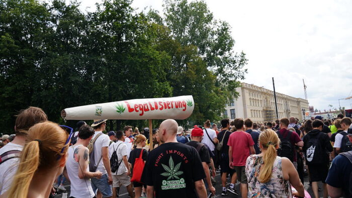 Viele Menschen demonstrieren auf einer Straße in Berlin, die gesäumt ist von Bäumen für die Legalisierung von Cannabis. Einige Demonstrierende halten einen gebastelten Joint hoch, der größer ist als ein Mensch. Darauf steht Legalisierung in roter Schrift. Links und rechts von dem Schriftzug sind grüne Hanfblätter zu sehen.