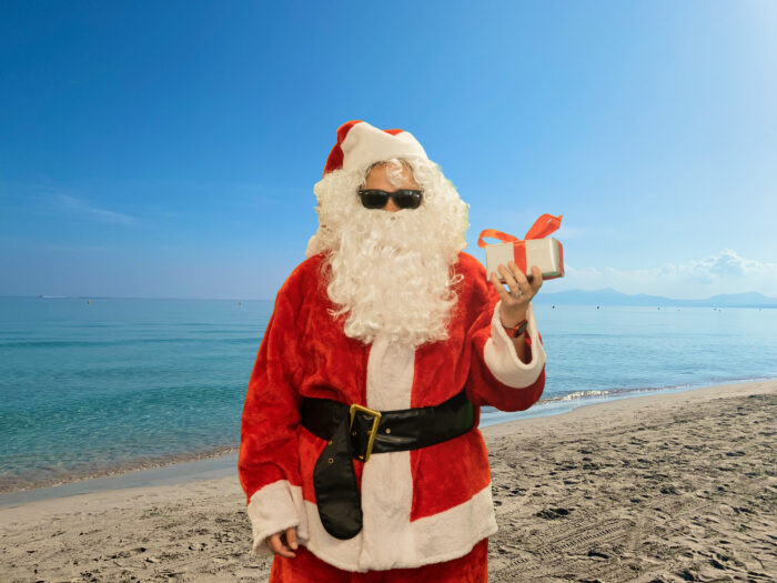 Ein als Weihnachtsmann verkleideter Mensch steht am Strand und hält ein kleines Päckchen in der Hand. Er trägt eine dunkle Sonnenbrille