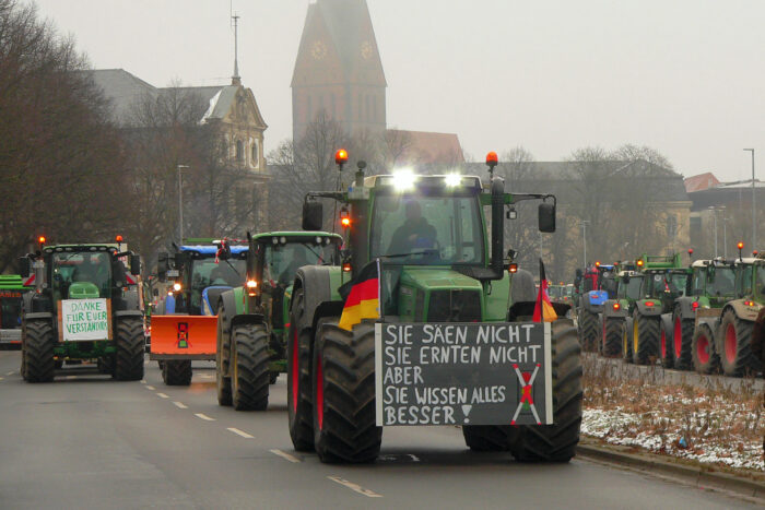 Eine Reihe von Traktoren mit Protestplakaten; auf dem ersten steht "Sie säen nicht, sie ernten nicht, aber sie wissen alles besser". Am Frontlader des Traktors sieht man zudem zwei Deutschland-Fahnen.