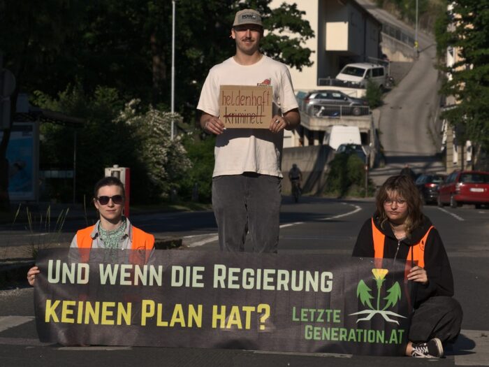 Zwei Aktivist*innen der Letzten Generation sitzen mit einem Transparent (Aufschrift: "Und wenn die Regierung keinen Plan hat?") auf der Straße. Zwischen ihnen steht Julian Schütter. Er hält ein Schild mit der Aufschrift "Heldenhaft" und dem durgestrichenen Wort "kriminell" in der Hand.