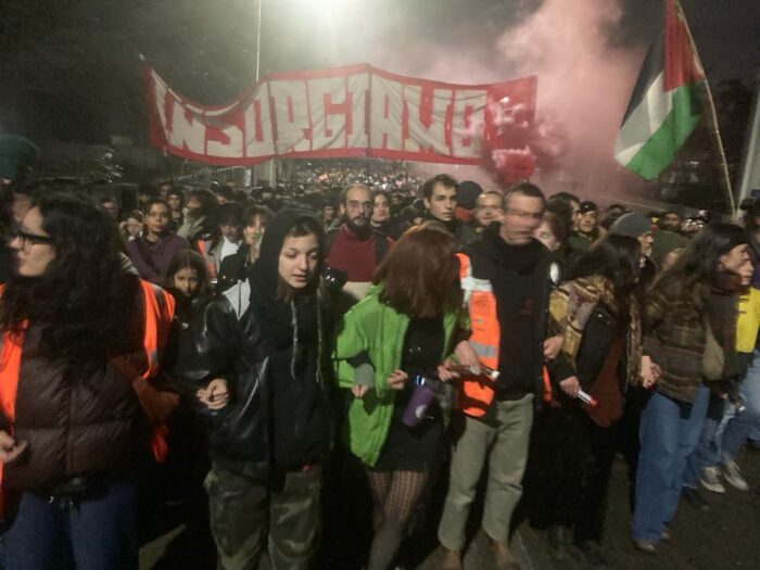 Mehrere Menschen haben sich eingehakt und bilden die erste Reihe einer Demonstration, für den Erhalt der besetzten Automobilteilefabrik GKN bei Florence/Italien. Einige Reihen weiter hinten halten Menschen ein Strangentrasparent in die Höhe, mit einer italinischen Aufschrift.