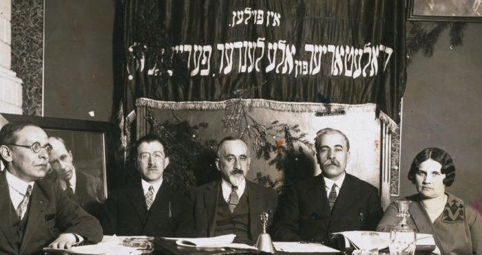 Eine Gruppe Personen sitzt im Anzug an einem Tisch, hinter ihnen ein festliches Banner mit hebräischen Buchstaben.