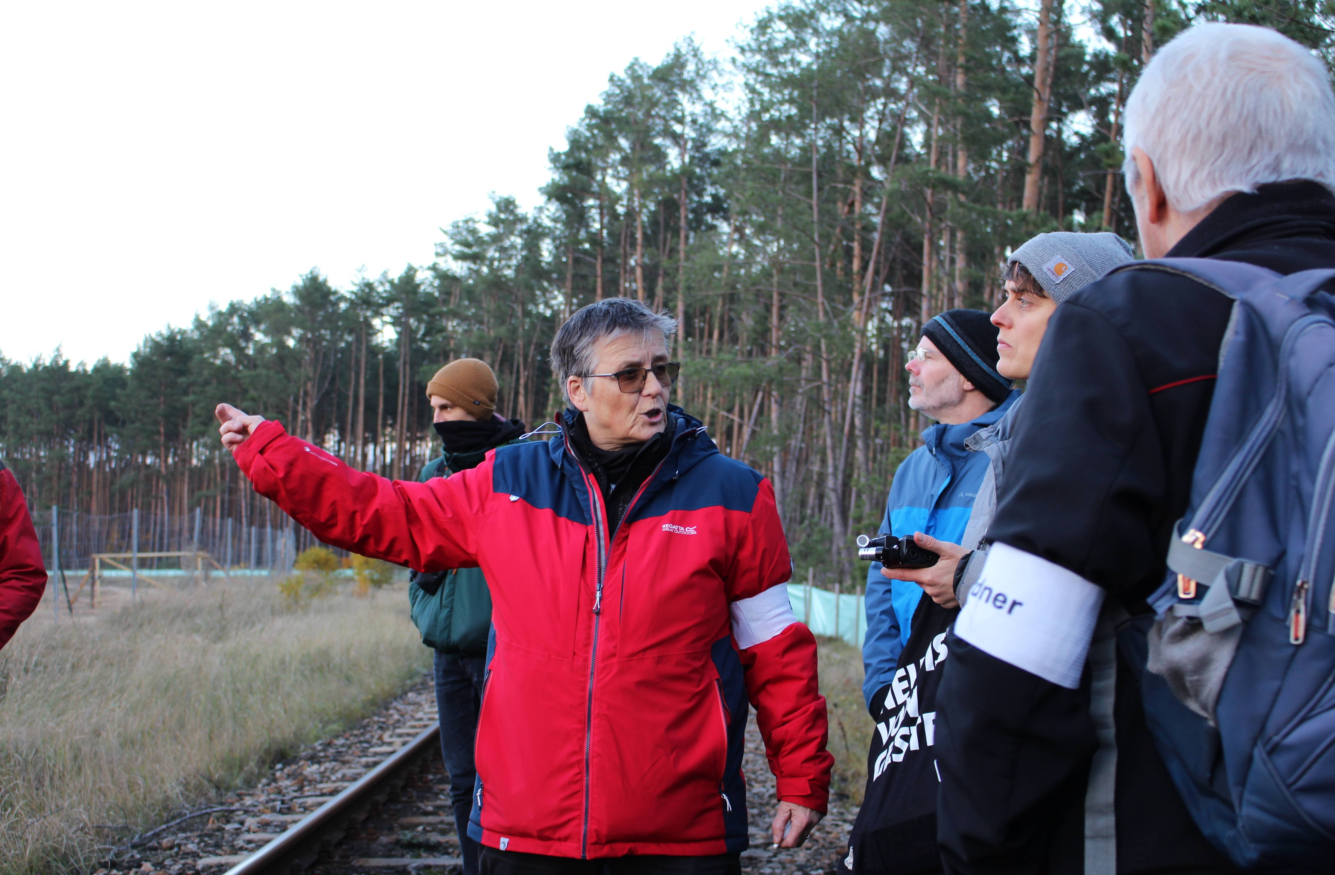 Eine mittelalte Frau mit kukrzen Haaren, Sonnenbrille und einer roten Outdoor-Jacke steht an einem Gleis an einem Waldstück und erklärt mehreren Zuhörer*innen etwas, während sie auf den linken Bildrand zeigt