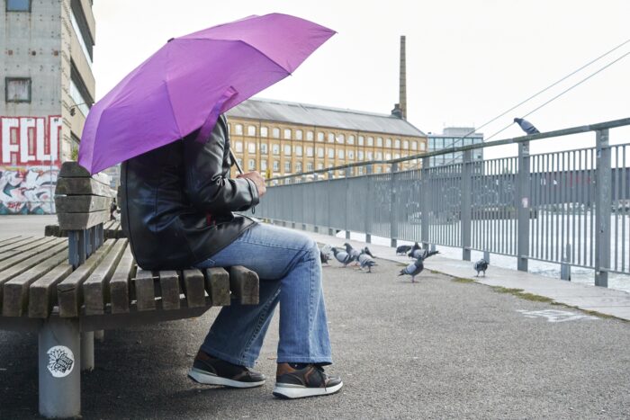 Eine Person in Lederjacke und Jeans und mit Regenschirm, der den Kopf verdeckt, sitzt an einem trüben Tag auf einer Bank und schaut aufs Wasser. Auf dem Geländer zum Wasser und auf dem Wegstück davor sitzen viele Tauben