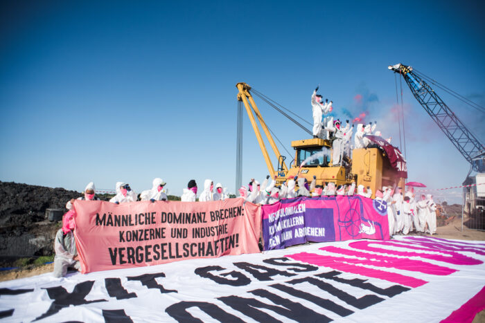 In weißen Overalls gekleidete Menschen, die einen Bagger besetzen und daneben ein Transparent hochhalten.