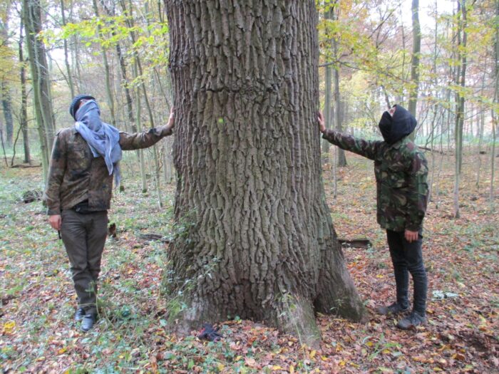 Zwei vermummte Menschen stehen links und rechts neben einem Baum und halten einen ausgestreckten Arm an ihn.