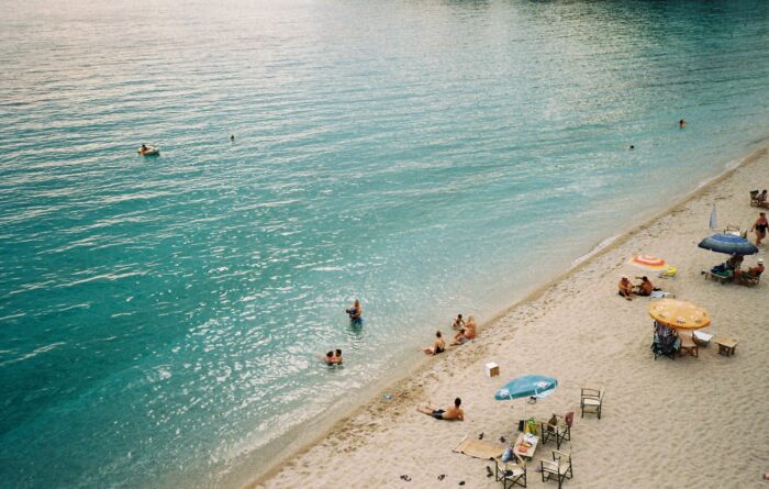 Blick von oben auf einen Strandabschnitt im Abendlicht. Am Strand sitzen mehrere Menschen, teils an der Wasserkante, Sonnenschirme sind zu sehen, ein paar Menschen schwimmen im Meer