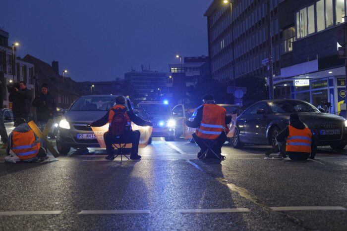 Eine Verkehrsblockade in orangenen Warnwesten der Letzten Generation, von hinten fotografiert, auf der Straße ist es noch Dunkel, man sieht Autoscheinwerfer und einen Polizeiwagen mit Blaulicht