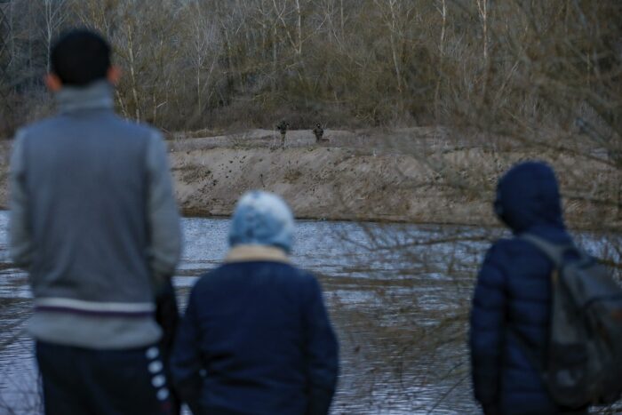 Drei junge Geflüchtete blicken von der türkischen Seite aus über den Evros und auf das andere Ufer. Auf der griechischen Seite sind zwei Soldat*innen zu sehen, die am Fluss patrouliieren. Das Bild wurde aus der Perspektive hinter den den drei Geflüchteten aufgenommen, man sieht sie von hinten