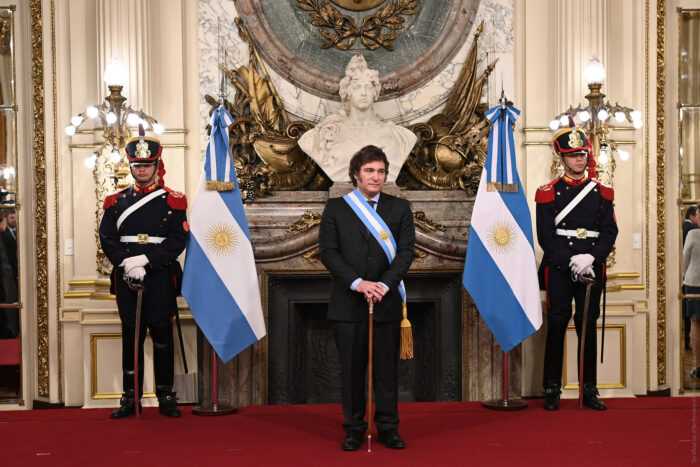 Der neue argentinische Präsident Javier Milei ist bei seiner Amtseinführung im Dezember 2023 zu sehen. Er steht vor einem Gemälde, in Anzug und mit einer Scherpe und hält einen Stock in der Hand. neben ihm stehen zwei argentinische Flaggen, daneben zwei Männer in traditionellen Wächter-Uniformen.