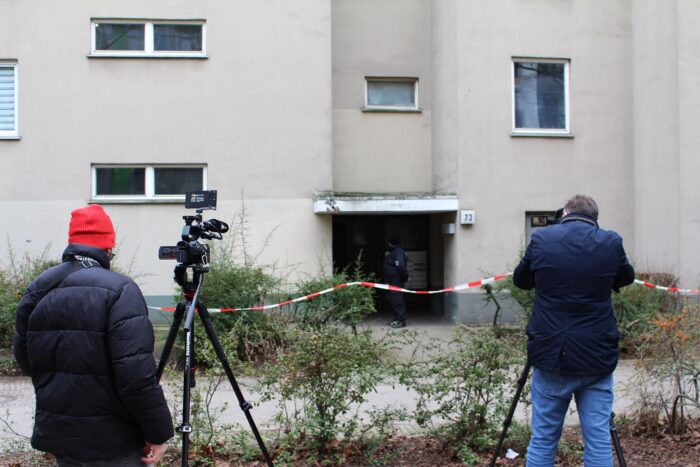 Fotografen stehen vor einem Hauseingang (ein grauer Neubau, Hausnummer 73), in einem dürren Strauch hängt Absperrband. Im Türbereich steht ein Polizist.