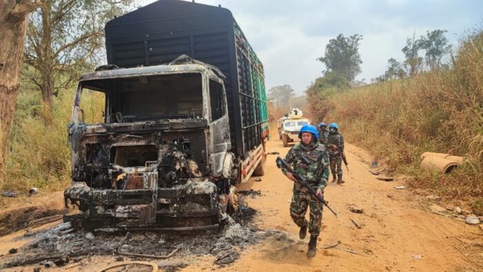 Aufnahme eines verbrannten LKW. An ihm vorbei laufen in einer Reihe Blauhelm-Soldaten, im Hintergrund fahren weiße Militärfahrzeuge