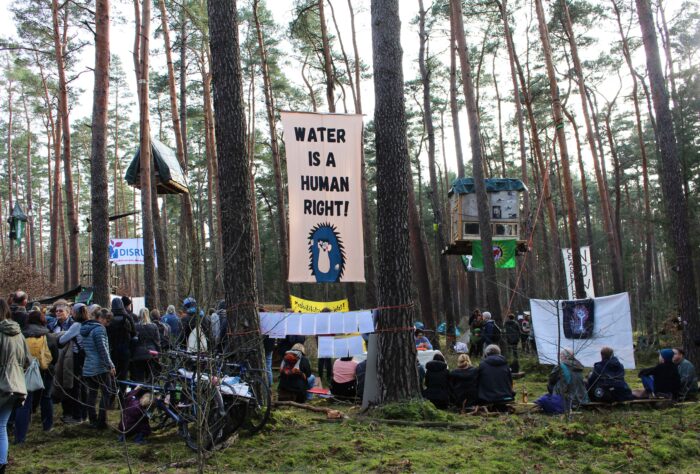 Menschenansammlung in einem Wald. Zwischen den Bäumen hängen Baumhäuser, von den Bäumen hängen Transparente. Auf dem vorderen ist ein Igel aufgemalt, darüber steht "Water is a human right".,