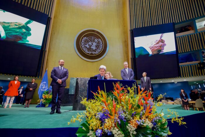 An einem Tisch unterschreibt John Kerry mit einem kleinen Mädchen auf dem Schoß ein Dokument. Links und rechts stehen zwei Männer in feierlicher Pose.