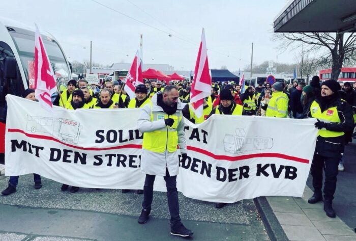 Grauer Tag an einem Busbahnhof. Eine Gruppe in ver.,di-Westen und m it ver.di-Gahnen hält ein Transparent mit der Aufschrift "Solidarität mit den Streikenden der KVB".