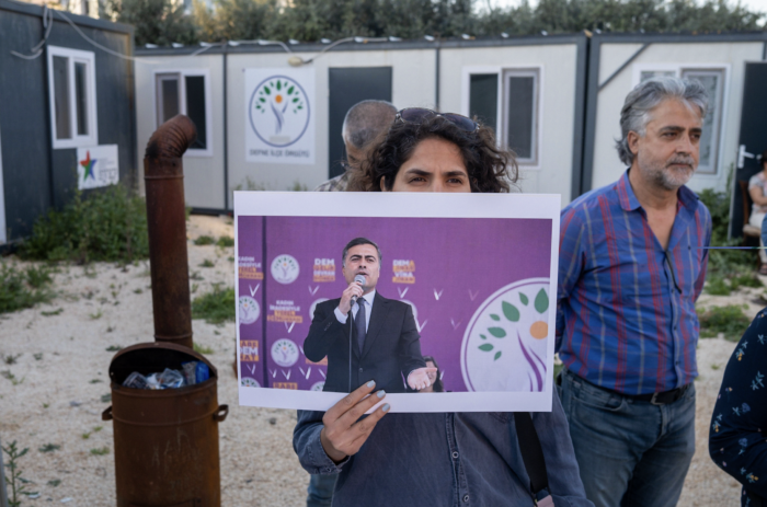 Eine Person steht vor Containergebäuden, an denen Banner der DEM Partei hängen. Die Person hält ein Schild mit dem Bild des gewählten Bürgermeisters von VAN hoch.