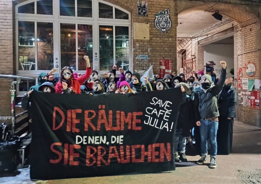 Aktivist*innen stehen mit erhobenen Fäusten hinter einem Banner mit der Aufschrift "Die Räume denen, die sie brauchen / Save Café Julia!"