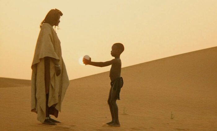 Filmstill aus »Yeelen«, in der Wüste übergibt ein kleiner junge einer Erwachsenen Person ein Ei, das von der dahinterstehenden Sonne erhellt wird.