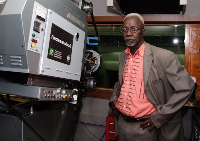Souleymane Cissé im schicken Anzug steht neben einem Filmprojektor und schaut in die Kamera