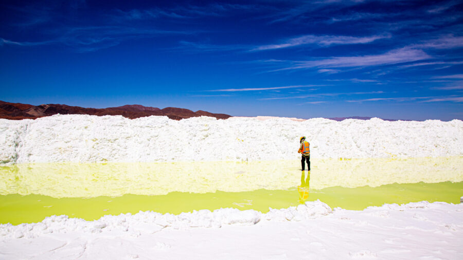 Ein Arbeite mit Helm und Signalweste steht in einem Becken mit einer griftgrünen Flüssigkeit. Drumherum ein großer Berg von getrocknetem Lithium, das aussieht wie weißer Pulver.