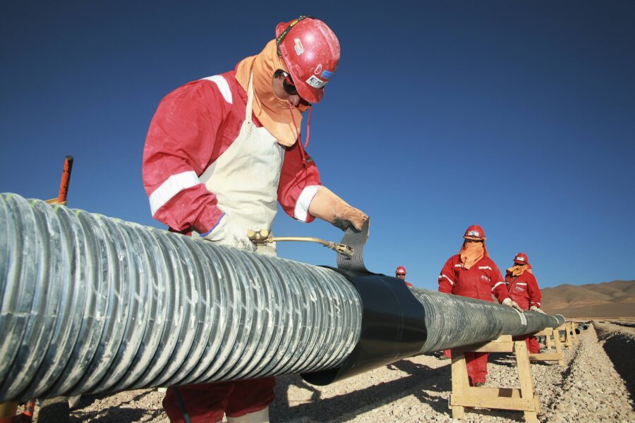 Bild von Arbeitern mit Helmen, signalroten Overalls, Sonnenbrille und vermummten Gesichtern. Ein Arbeiter verschweißt metallische Röhren.