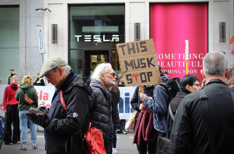 Mehrere Menschen stehen vor einer Tesla Filiale, eine Person hält ein Schild hoch, auf dem steht "This Musk stop"