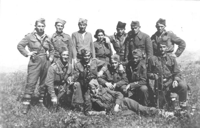 Auf dem Bild sind 13 Partisan*innen zu sehen, die auf einer Wiese posieren. Sie tragen Uniformen und Barette. Manche haben eine Waffe in der Hand.