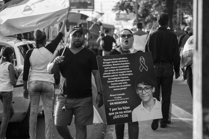 Bild einer Demonstration. Im Mittelpunkt zwei Protestierende, eine hält ein Trauerplakat mit einem Text und dem Bild eines jungen Mannes mit Brille