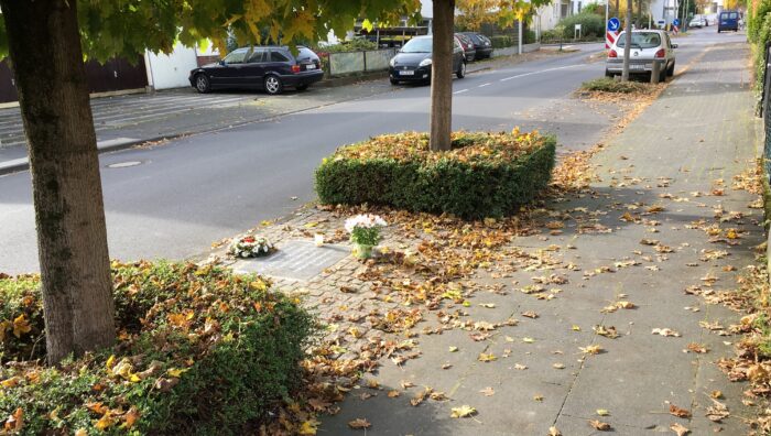Auf einem Gehweg, zwischen zwei Bäumen, ein Blumenkranz und eine Gedenktafel und Laub.