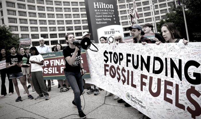 Eine Frau mit Megafon vor einem Transparent mit der Aufschrift "Stop Funding Fossil Fuels"