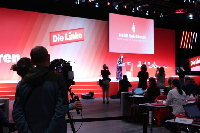 Eine große Rote Bühne mit Schriftzug "Die Linke". Vor der Bühne sind ein paar Stuhlreihen zu sehen, im Vordergrund ein Kamerateam, direkt vor dem Podium ebenfalls mehrere Fotograf*innen. Auf der Bühne spricht Heidi Reichinnek am Rednerpult