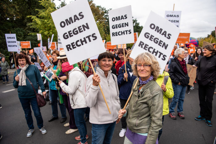 Das Bild zeigt eine Demo von älteren weiblich gelesenen Personen, die Schilder in Höhe halten mit der Aufschrift: Omas gegen Rechts. Zwei stehen der Kamera zugewandt und lächeln.