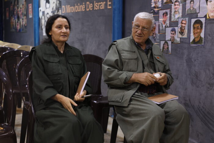 Besê Hozat und Cemîl Bayik sitzen auf Stühlen beim PKK-Kongress. Besê Hozat spricht und hält einen Notizblock und einen Stift in der Hand. Cemîl Bayik blickt neben ihr lächelnd in den Raum. Im Hintergrund sind weitere Stühle und Bilder von gefallenen Kämpfer*innen an der Wand.
