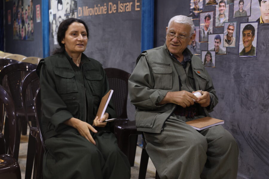 Besê Hozat und Cemîl Bayik sitzen auf Stühlen beim PKK-Kongress. Besê Hozat spricht und hält einen Notizblock und einen Stift in der Hand. Cemîl Bayik blickt neben ihr lächelnd in den Raum. Im Hintergrund sind weitere Stühle und Bilder von gefallenen Kämpfer*innen an der Wand.