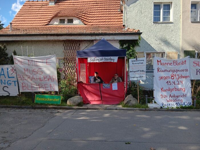 Man sieht einen roten Pavillon, in dem zwei ältere Menschen sitzen, umgeben von Transparenten.