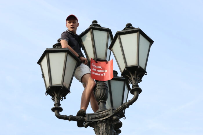 Man sieht eine Person, die auf einer Straßenlaterne sitzt und ein rotes Schild hält, darauf steht in kyrillischen Buchstaben auf Russisch: Putin, wo ist user Geld?