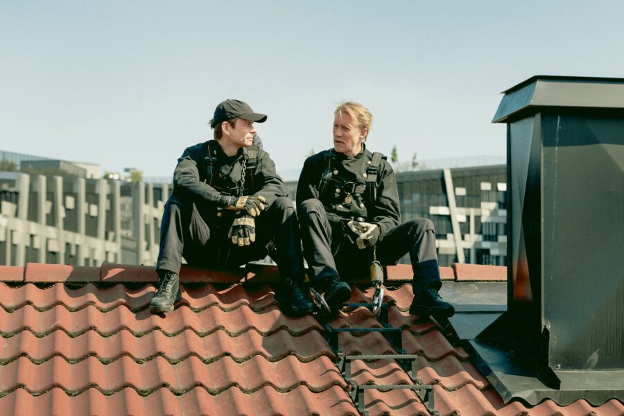 Zwei Schornsteinfeger sitzen auf einem Dach und unterhalten sich. Der Himmer ist Wolkenlos. Einer hat dunkle Haare und trägt eine Cappy. Der andere hat blonde Haare und einen Schnauzbart. Beide haben Sicherheitskleidung an. Neben ihnen ist ein großer schwarzer Schornstein.