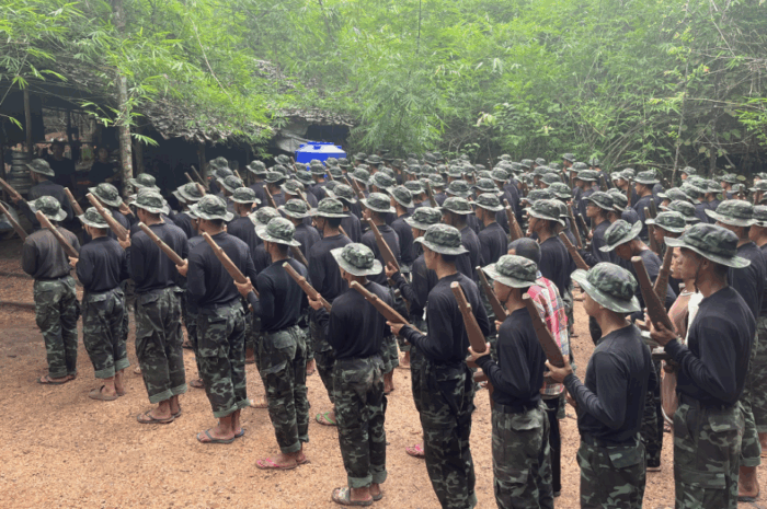 DAs Bild zeigt bewaffnete Kämpfer*innen in Myanmar die in mehreren Reihen stehen. Sie tragen Militärkleidung und Gewehre im Anschlag. Sie stehen auf einem stabigen Platz, mit Bäumen im Hintergrund